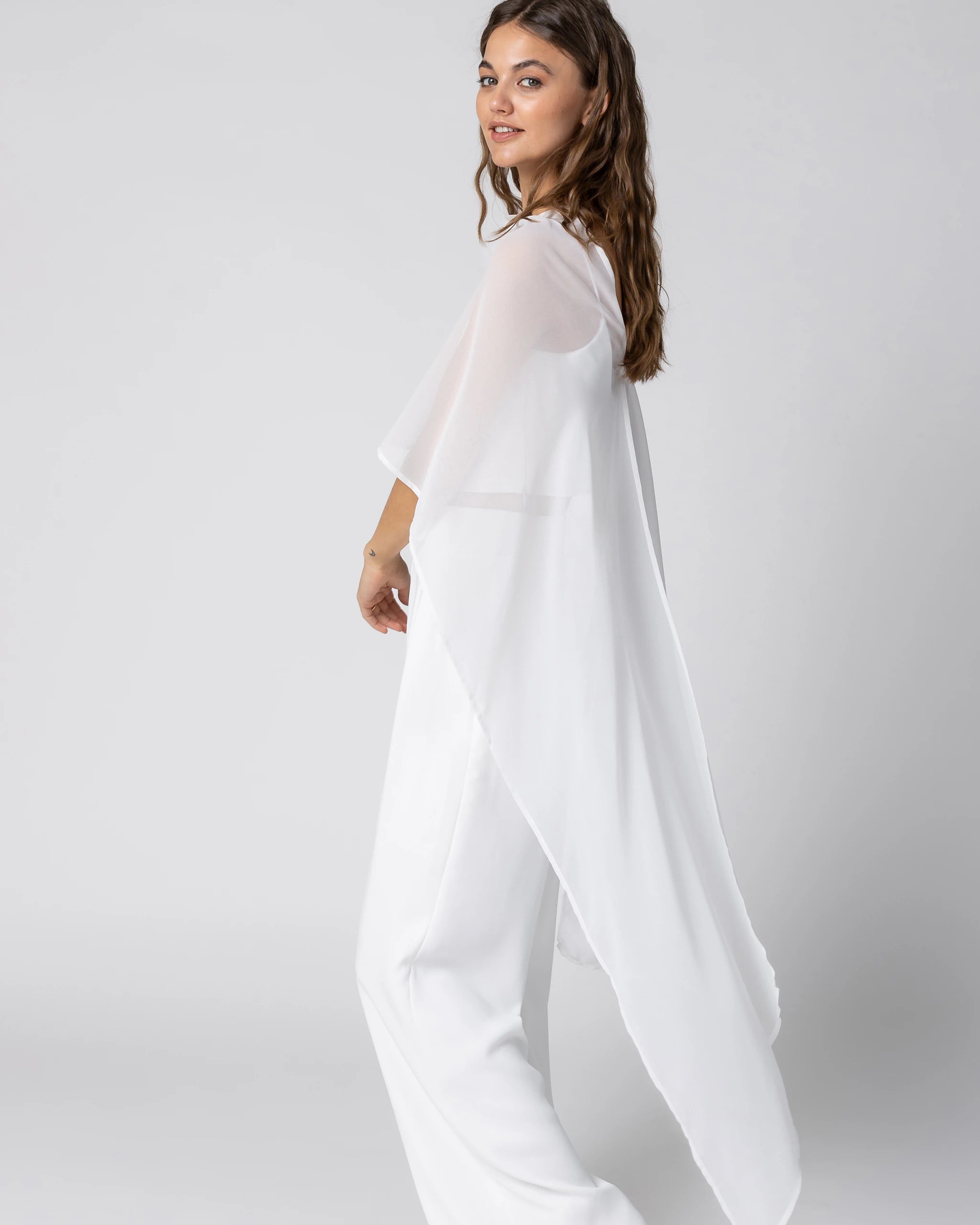Braut in modernem Braut Jumpsuit mit Bralette Top, weiter Marlene Hose und Chiffon Cape mit Blütenbrosche Ansicht seitlich