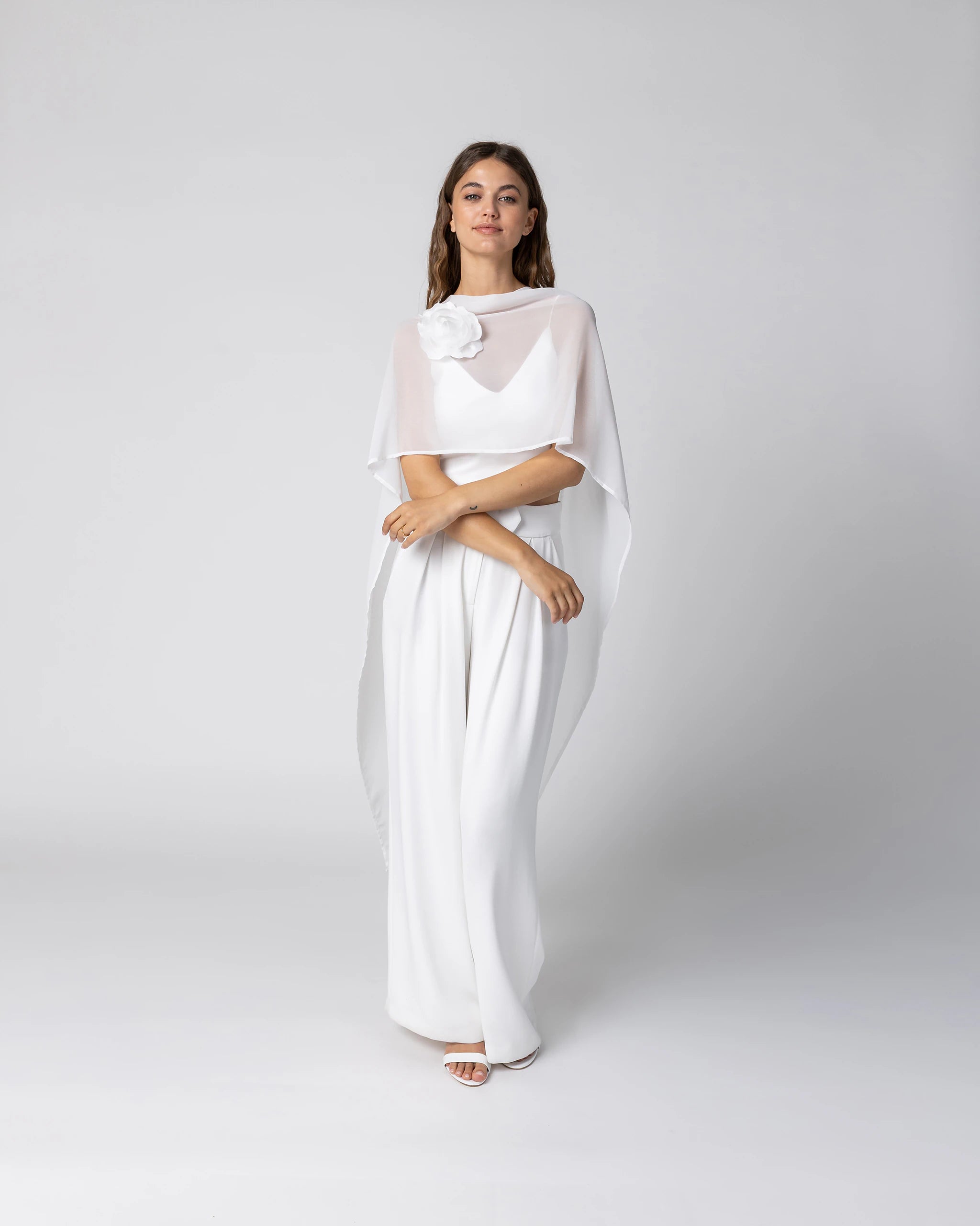 Braut in modernem Braut Jumpsuit mit Bralette Top, weiter Marlene Hose und Chiffon Cape mit Blütenbrosche Ansicht vorne